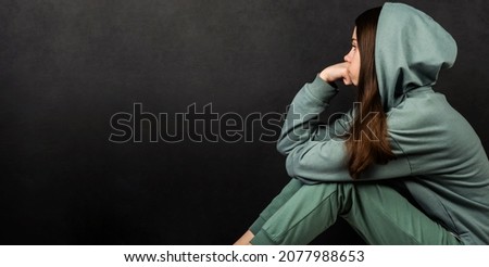 A teenage girl in a green sweatshirt sits alone and is sad against a dark background. Adolescent psychology concept. Emotions. Royalty-Free Stock Photo #2077988653