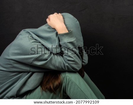 Teenage girl in a green sweatshirt covers her face with her hands on a dark background. The girl sits alone and is afraid. Adolescent psychology concept. Emotions. Royalty-Free Stock Photo #2077992109