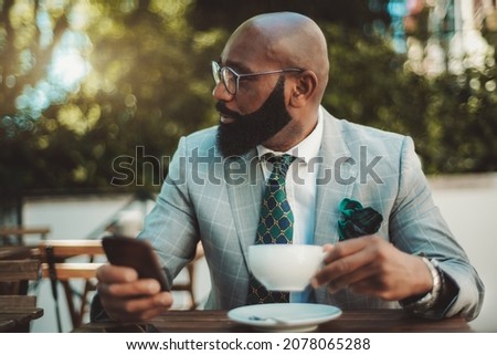 A portrait of a handsome dapper bald mature black man entrepreneur with a well-groomed beard and in a formal suit with a necktie, sitting in a street restaurant and drinking tea during a coffee break Royalty-Free Stock Photo #2078065288