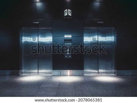 A dark hall of an airport lit by a bluish neon glow with two modern elevators in the middle with chromium closed doors and spots of light near each lift and a control panel
