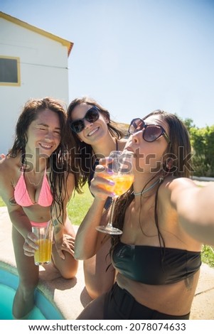 Pleased female friends taking selfie at poolside. Women in swimsuits resting in swimming pool, taking pictures with mobile phone. Leisure, friendship, party concept