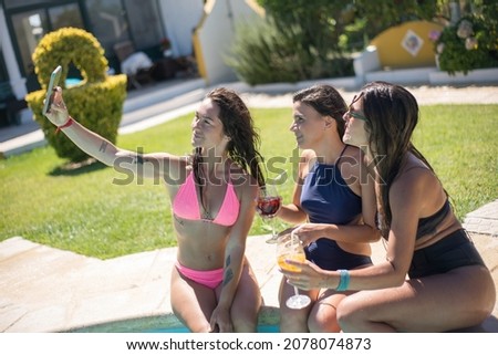 Female friends taking selfie at poolside. Women in swimsuits resting in swimming pool, taking pictures with mobile phone. Leisure, friendship, party concept