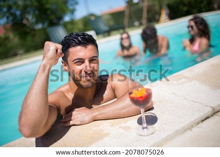 Sporty man and bright cocktail in swimming pool. Man with dark hair and glass with bright beverage, looking at camera, posing. Leisure, friendship, party concept