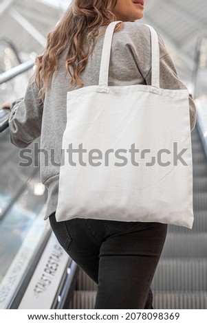 Young woman using white blank Tote bag posing in mall. Urban minimal clothing product apparel style business. Photo template for mockup, print store, e-commerce shop, branding design project.