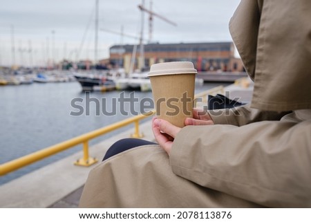 Girl drinks coffee or tea on the background of yachts in the Marina. Hot drink in a paper cup. Disposable paper cup close up. Empty space for text, layout