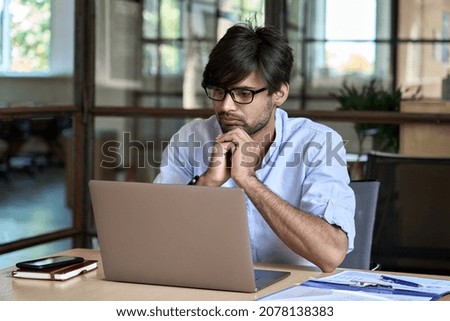 Young Indian businessman using computer working online watching business training webinar in office. Ethnic guy employee thinking on project analysis looking at laptop at workplace. Royalty-Free Stock Photo #2078138383
