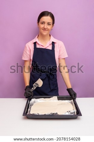 Woman in an apron and gloves holds baking sheet with dough, which lies on table and looks at camera. To advertise recipes. Selective focus.