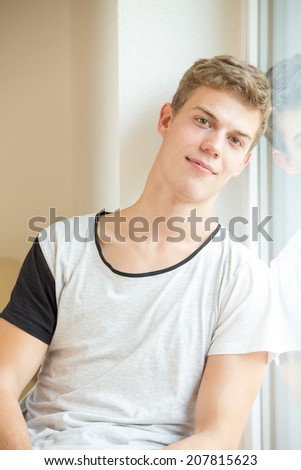 A young male model is smiling into the camera while leaning on a window - mouth closed