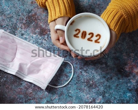 Hello 2022 Goodbye COVID-19, holidays food art theme female hands holding coffee cup with number 2022 over frothy surface on rustic blue background with disposable medical mask. (selective focus)