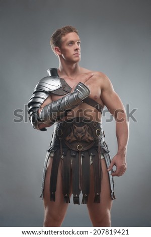 Half length portrait of young handsome muscular man gladiator in armour pointing up isolated over grey background
