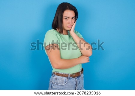 Very bored Young european girl wearing green T-shirt over blue background holding hand on cheek while support it with another crossed hand, looking tired and sick,