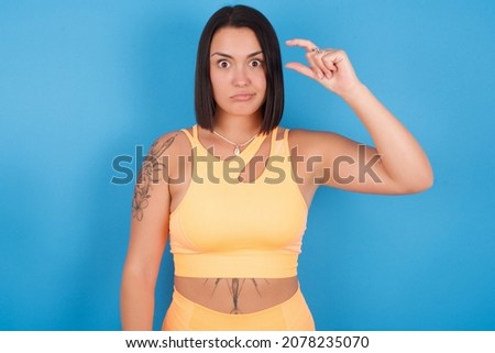 Young caucasian sporty girl standing against blue background purses lip and gestures with hand, shows something very little.