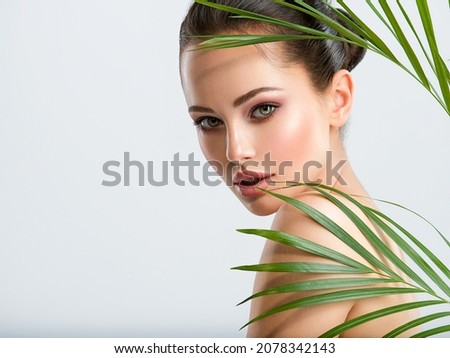 Young beautiful woman with healthy skin of face and palm leaves. Closeup fresh face of an attractive caucasian girl with green plants. Model with bright brown eye makeup. Skin care concept. 