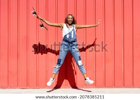 Black woman dressed casual, wtih a skateboard jumping for joyon red urban wall background.