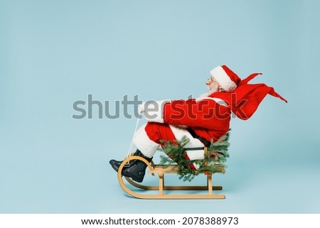 Full length side view old Santa Claus man 50s in Christmas hat red suit coat clothes sitting in sled isolated on plain blue background studio. Happy New Year 2022 celebration merry ho x-mas concept