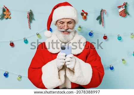 Old bearded Santa Claus man 50s wears Christmas hat red suit clothes hold use mobile cell phone isolated on plain blue background studio. Happy New Year 2022 celebration merry ho x-mas holiday concept