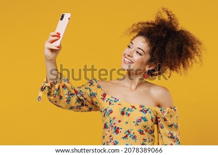 Young happy smiling beautiful woman 20s with culry hair wearing casual clothes doing selfie shot on mobile cell phone post photo on social network isolated on plain yellow background studio portrait.