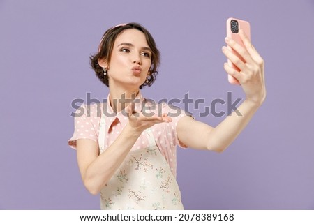 Young housewife housekeeper chef cook baker woman in pink apron doing selfie shot on mobile phone post photo on social network blow air kiss isolated on pastel violet background. Cooking food concept.
