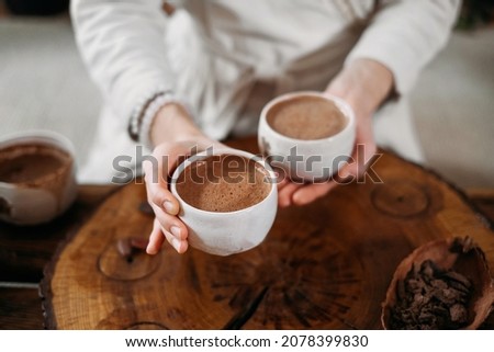 Hot handmade ceremonial cacao in white cup. Woman hands holding craft cocoa, top view on wooden table. Organic healthy chocolate drink prepared from beans, no sugar. Giving cup on ceremony, cozy cafe Royalty-Free Stock Photo #2078399830