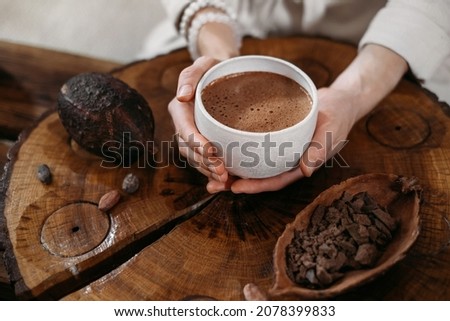 Hot handmade ceremonial cacao in white cup. Woman hands holding craft cocoa, top view on wooden table. Organic healthy chocolate drink prepared from beans, no sugar. Giving cup on ceremony, cozy cafe Royalty-Free Stock Photo #2078399833