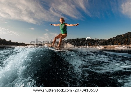 happy woman energetically balancing on wave on wakesurf board at sunny day. Blue sky and trees in the background.