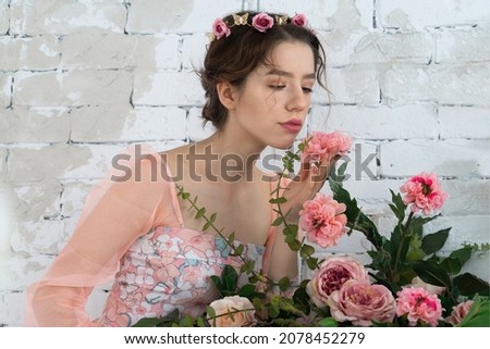 A tender young girl in the image of a princess in a pink dress with a wreath on her head sniffs a bouquet of red and pink roses.