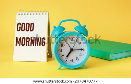 Clock and notepad with the text Good morning on a yellow background, top view.