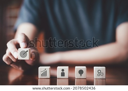 Businessman holding wooden cube with target board, chart, idea icon and on wooden table. Goals and planning for success in marketing business, achieve the objective concept. Closeup free copy space.