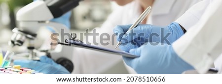 Woman chemist looking through microscope in laboratory. Assistant writing data to paper. Laboratory diagnostics of diseases concept Royalty-Free Stock Photo #2078510656
