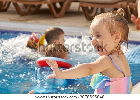 A little girl in rainbow swimsuit sits on the swimming pool and laughing in sun on the background of bathing children. Children recreation in the summer, near the pool.