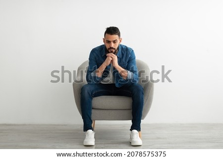 Negative emotions. Young Arab man sitting in armchair with dull or thoughtful face expression, feeling unhappy or tired, having problem, suffering from depression against white studio wall Royalty-Free Stock Photo #2078575375