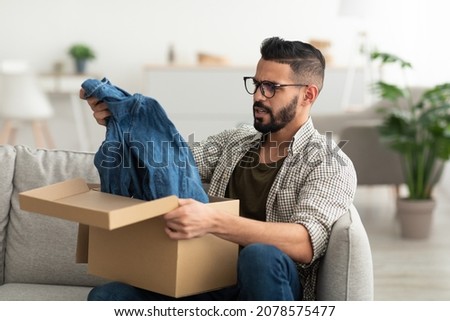 Online shopping mistake, terrible purchase, wrong delivery concept. Unhappy young Arab guy opening carton package, receiving clothes, dissatisfied with delivered item at home Royalty-Free Stock Photo #2078575477