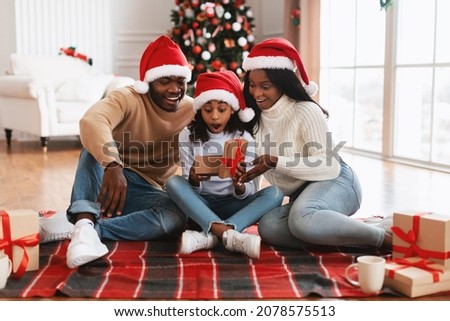 Wow. Cute excited black girl opening Christmas present box in festive living room. Smiling parents giving Xmas gift to surprised daughter sitting on floor blanket near decorated tree on holiday eve