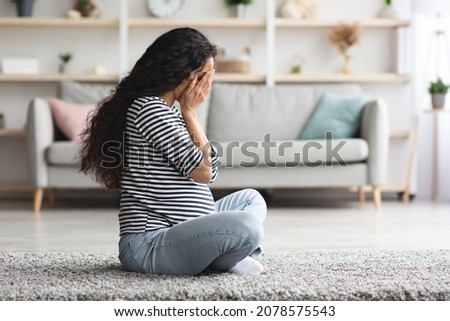 Pregnancy hormonal changes concept. Young curly long-haired pregnant woman feeling down, sitting on floor in cozy living room and crying, staying alone at home, side view, free space Royalty-Free Stock Photo #2078575543