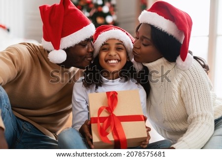 Portrait of cheerful African American parents kissing smiling excited daughter in cheeks, girl holding Xmas holiday gift box, happy people in red Santa Claus hats celebrating Christmas at home