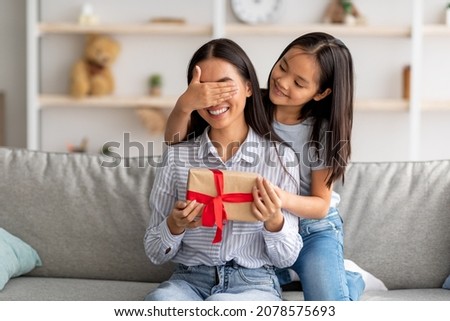 Playful asian girl making surprise for her happy mother, closing her eyes and giving gift box while sitting on couch at home interior, copy space