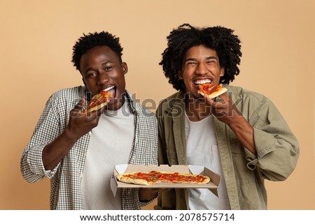 Tasty Snack. Two Hugry Black Guys Eating Italian Pizza From Box And Looking At Camera, Cheerful African American Male Friends Enjoying Food Delivery Service, Standing On Beige Background, Free Space