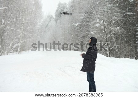 A man takes pictures on a quadcopter of a winter forest during a snowfall. He has a remote control from the drone in his hand. The aircraft hovered in the air.Selective focus.                         