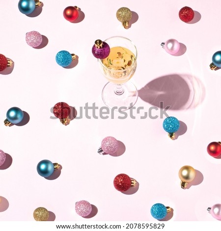 Glass of white wine decorated with a purple Christmas ball and many balls of different colors around and and their shadow. Minimal New Year’s concept on a pink background