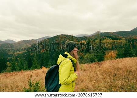 profile photo of a girl wearing warm professional hiking bright clothes walking in mountains covered with forest in autumn, cloudy sky