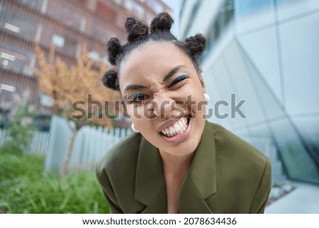 Close up shot of cheeky dark skinned woman winks eye clenches teeth wears blue eyeliner dressed in fashionable clothes poses in urban setting has fun. Positive teenager spends free time outdoors Royalty-Free Stock Photo #2078634436