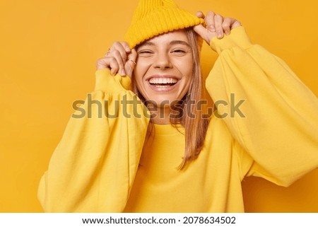 Happy playful young woman smiles broadly shows white teeth keeps hands on hat dressed in casual jumper expresses sincere emotions being in good mood isolated over yellow background. Joy concept Royalty-Free Stock Photo #2078634502