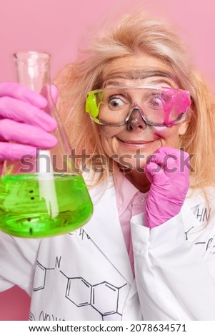 Vertical shot of surprised female scientist looks at green liquid in glassware has dirty glasses white medical coat isolated over pink background. Chemical studies and laboratory experiments concept