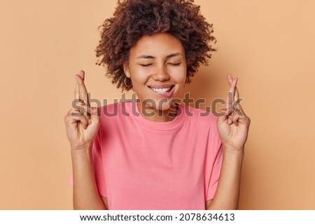 Photo of positive hopeful woman bites lips keeps fingers crossed prays and anticipates for something keeps eyes closed dressed in casual pink t shirt isolated over beige background makes wish