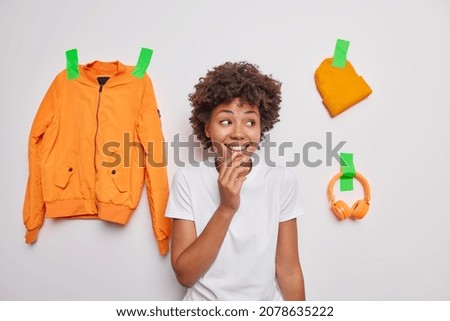 Glad curly haired young woman smiles happily notices something pleasant away dressed in casual t shirt poses against white background with orange jacket hat and headphones plastered to wall.