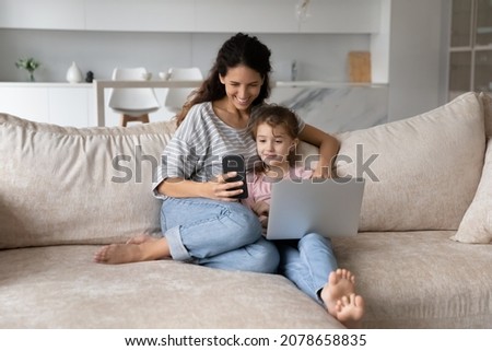 Happy mother with cute daughter having fun with electronic devices at home, using smartphone and laptop together, smiling mom and adorable girl kid chatting online by video call, watching cartoons
