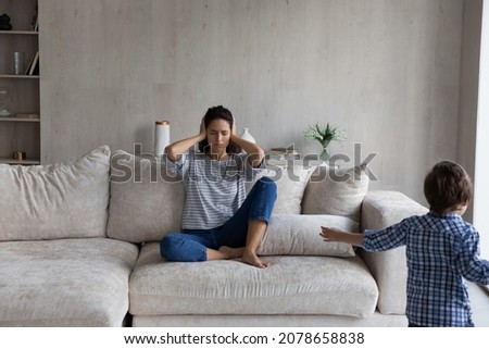 Young woman sit on sofa covers ears with palms feels tired due active noisy little son. Disobedient boy running in living room disturb the peace of mom. Upbringing problems, hyperactive child concept Royalty-Free Stock Photo #2078658838
