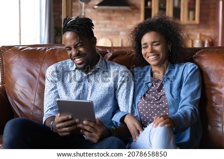 Joyful millennial African ethnicity family couple using digital tablet, laughing watching funny photo video content in social network, enjoying comedian movie. resting together on cozy sofa at home. Royalty-Free Stock Photo #2078658850