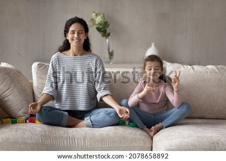 Happy mother and little daughter practicing yoga, meditating on comfortable couch at home together, smiling calm mom teaching cute girl kid to relaxing, doing exercises, family meditation concept