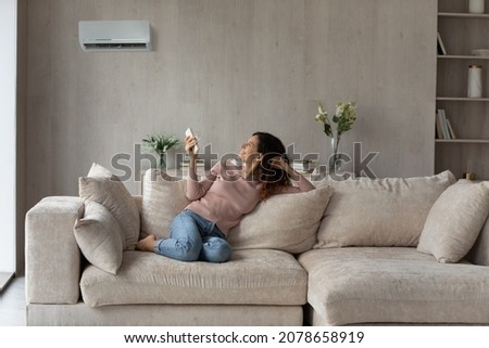 Smiling woman using air conditioner remote controller, relaxing on comfortable couch in living room alone, positive beautiful young female switching temperature on climate control system at home Royalty-Free Stock Photo #2078658919
